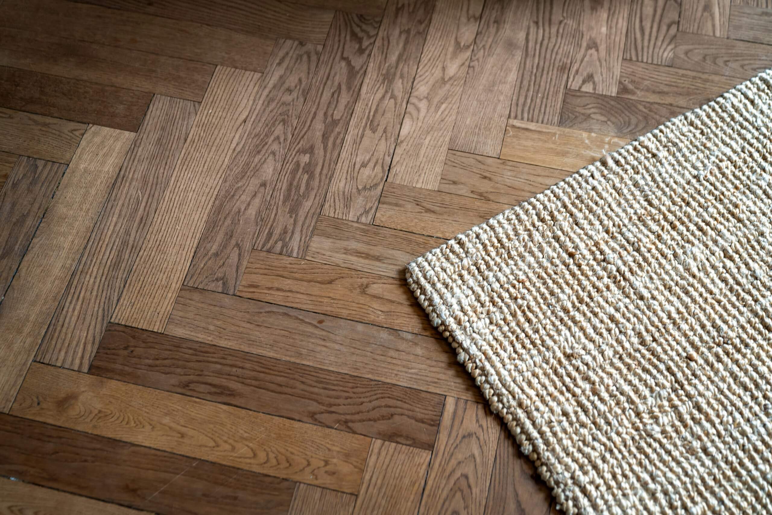 A beige woven rug lies on an eco-friendly flooring with a wooden herringbone pattern. The textures of the rug and wood contrast with each other.