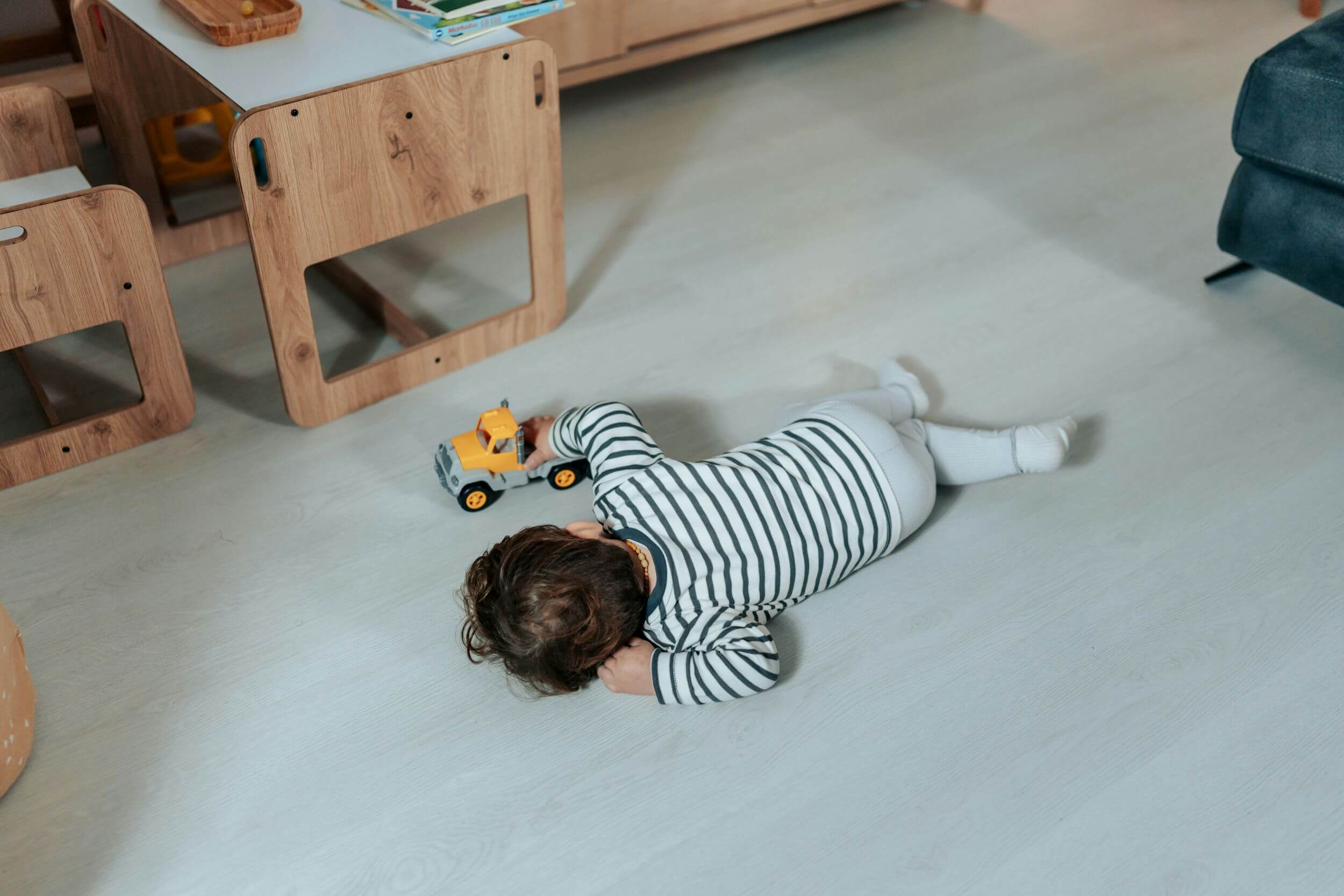A young child in a striped outfit lies on the floor, playing with a yellow toy truck in a cozy, light-colored room with kid-friendly flooring options and wooden furniture.