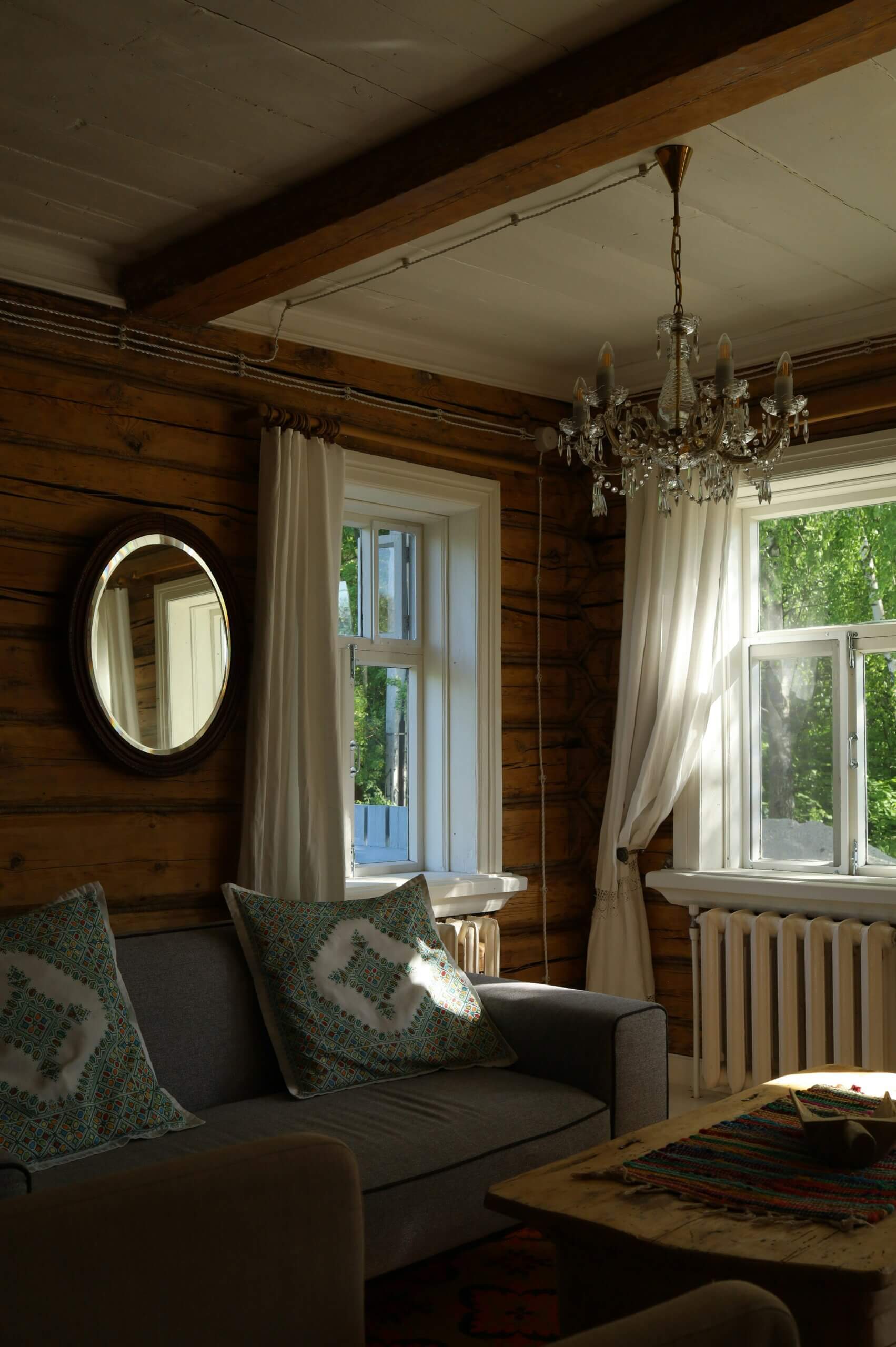 A cozy living room with wooden walls, a gray sofa and patterned pillows, white curtains framing two windows, a round mirror, elegant chandelier, soft carpet and flooring, and sunlight streaming in to create a warm, inviting atmosphere.