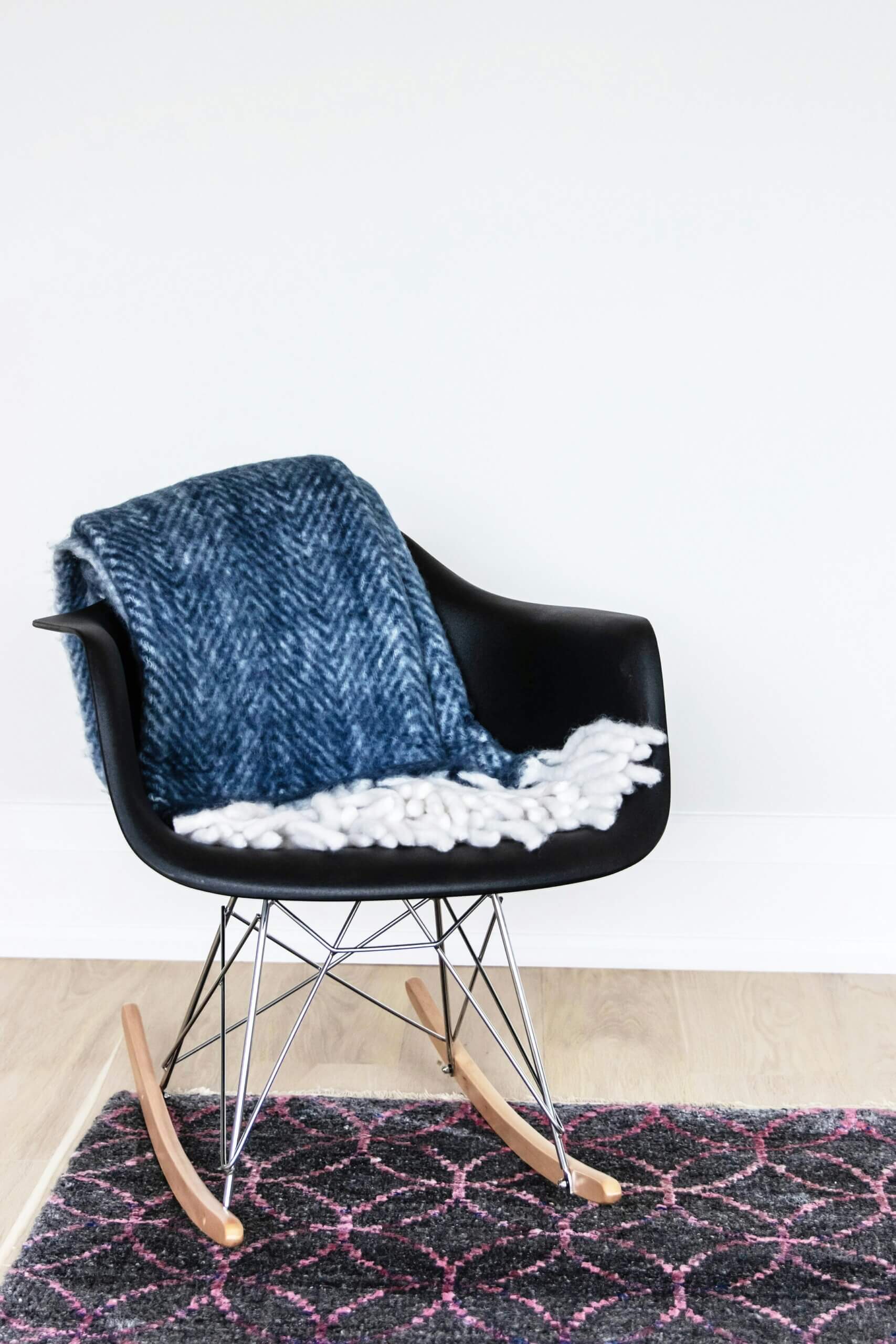 A modern black rocking chair with wooden rockers holds a folded blue patterned blanket and a white textured throw, placed on a rug with a pink and gray geometric design—perfect for showcasing your unique style and flooring tips against a white wall.