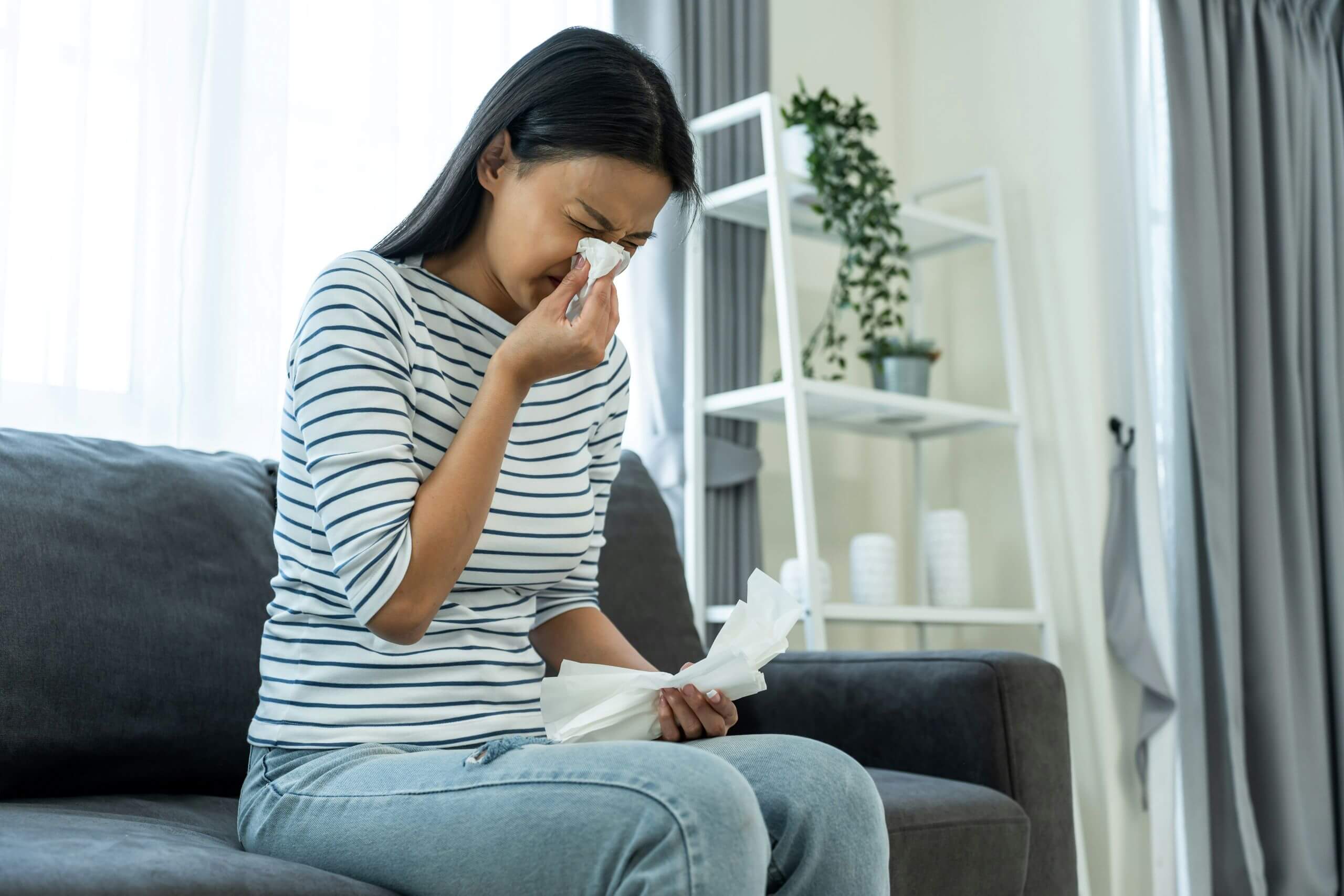 A woman sits on a couch, holding tissues and wiping her nose with one. She appears upset or unwell, in a bright living room with a shelf and potted plant in the background.
