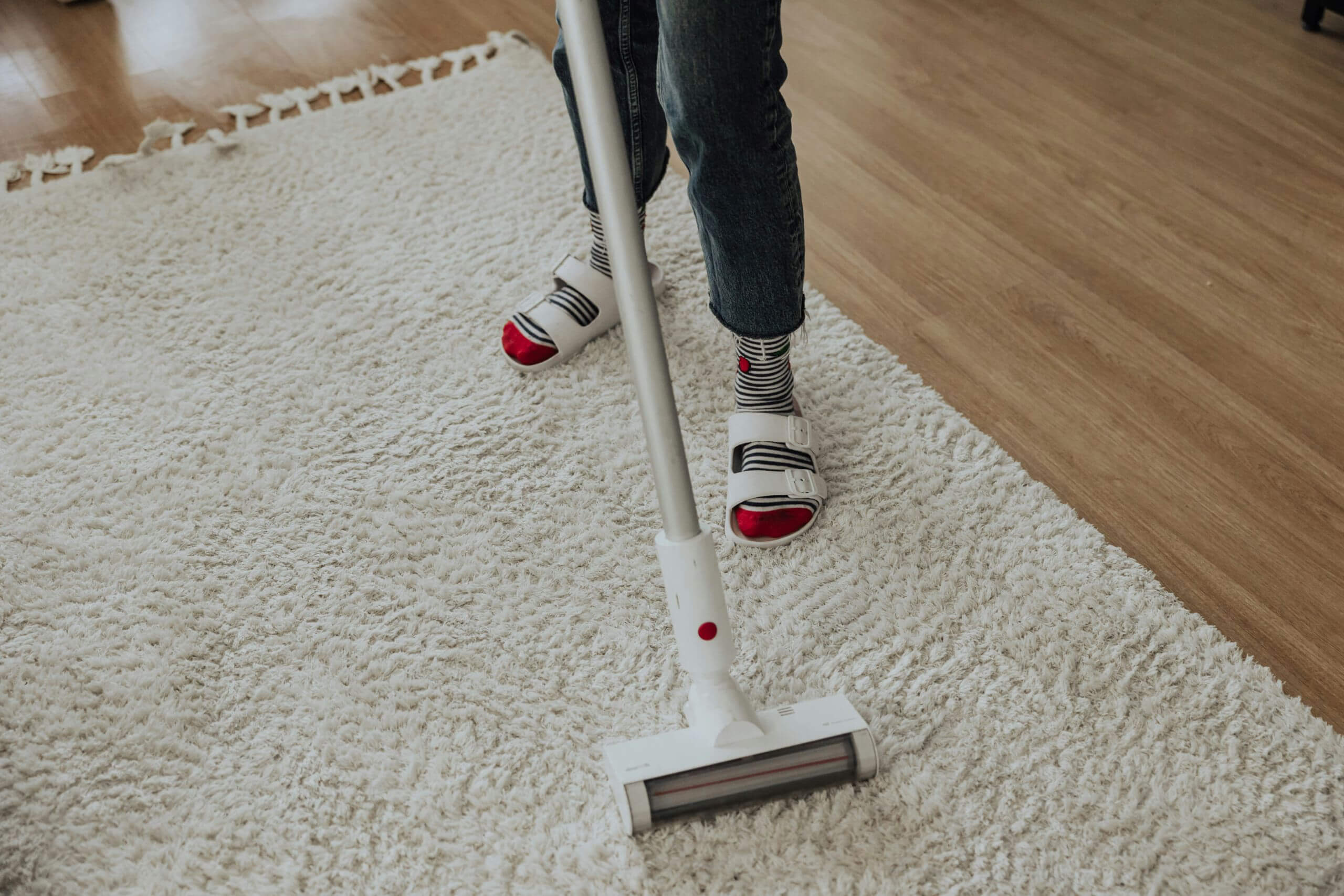 Person wearing striped socks and jeans using a cordless vacuum cleaner on a white, fluffy rug next to a wooden floor.