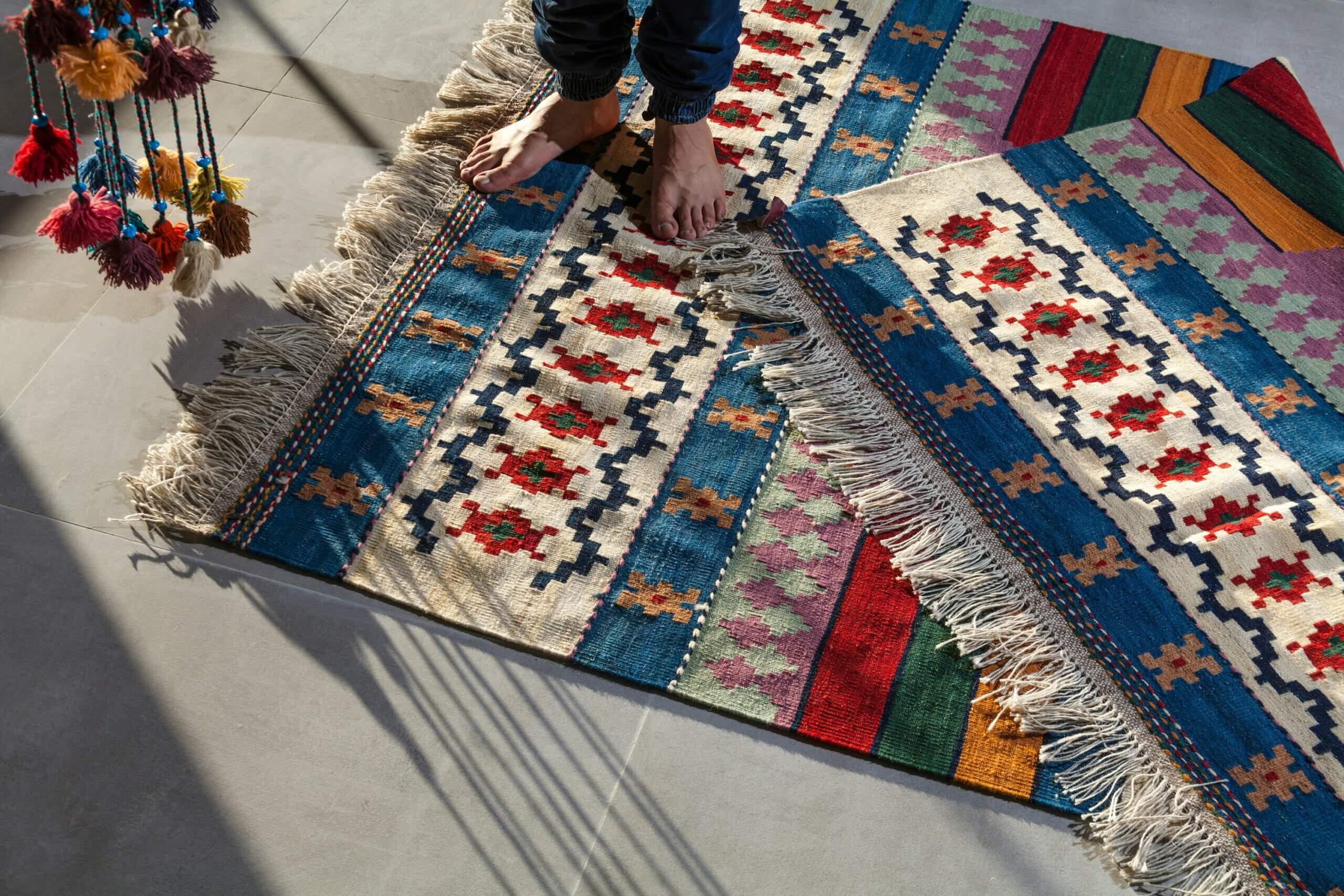 A person with bare feet stands on colorful, patterned rugs with fringed edges, illuminated by sunlight streaming through a window, casting shadows on a tiled floor.