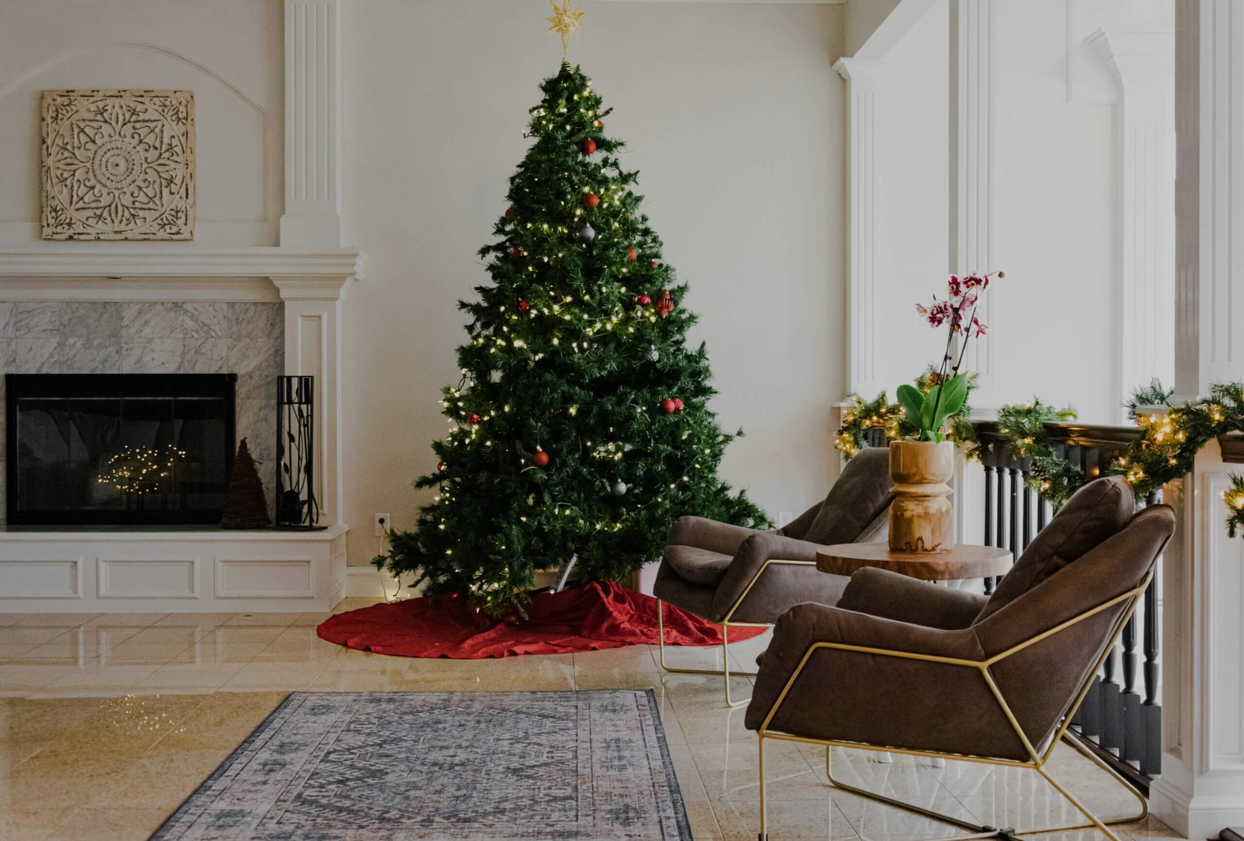 A decorated Christmas tree with lights and ornaments stands in a bright living room next to a fireplace. Two modern chairs and a small table sit nearby, with festive garlands on the railing.