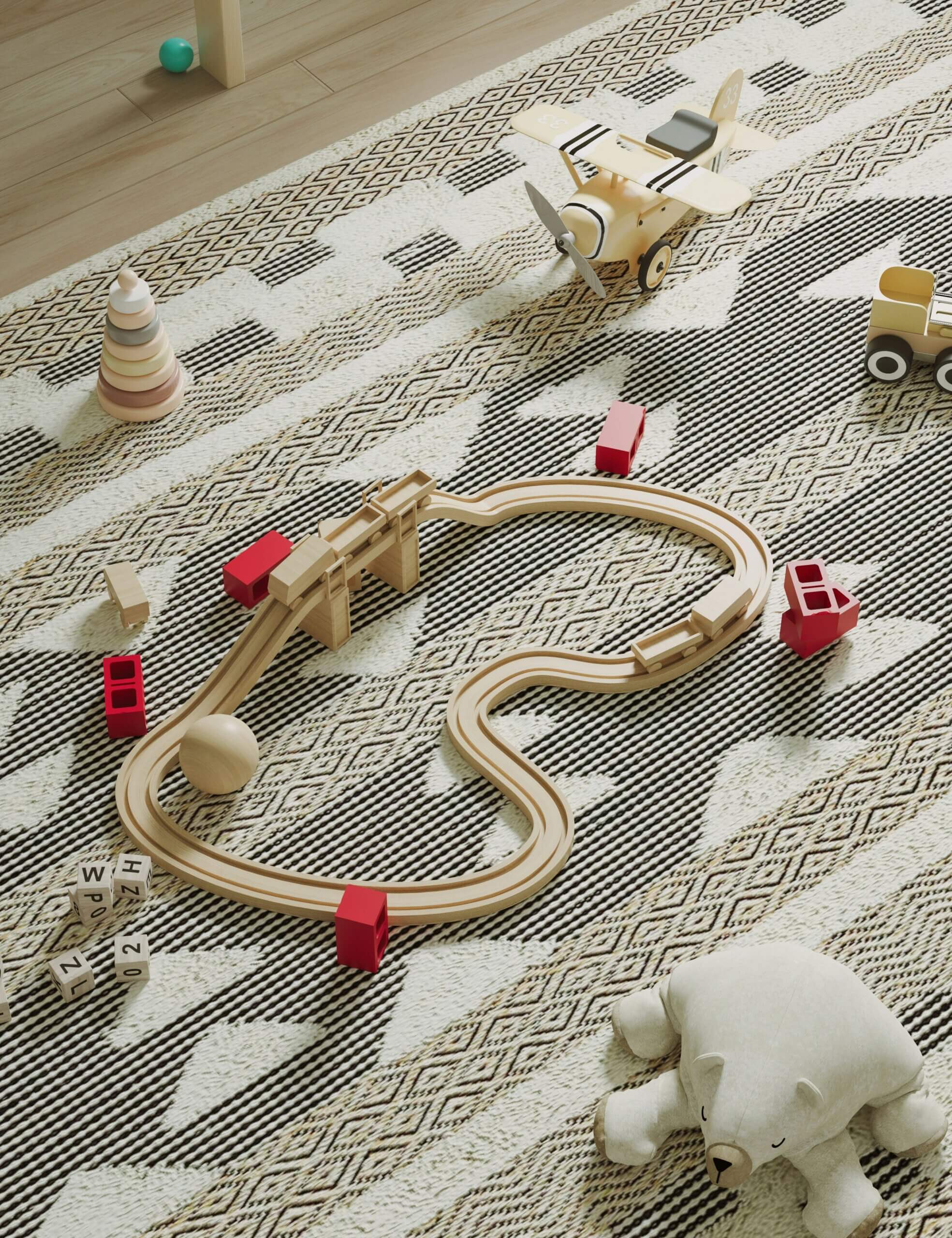 Wooden toy train tracks with red bridges, a wooden airplane, blocks, a stacking toy, and a stuffed bear are scattered on a black-and-white patterned rug in a playroom.