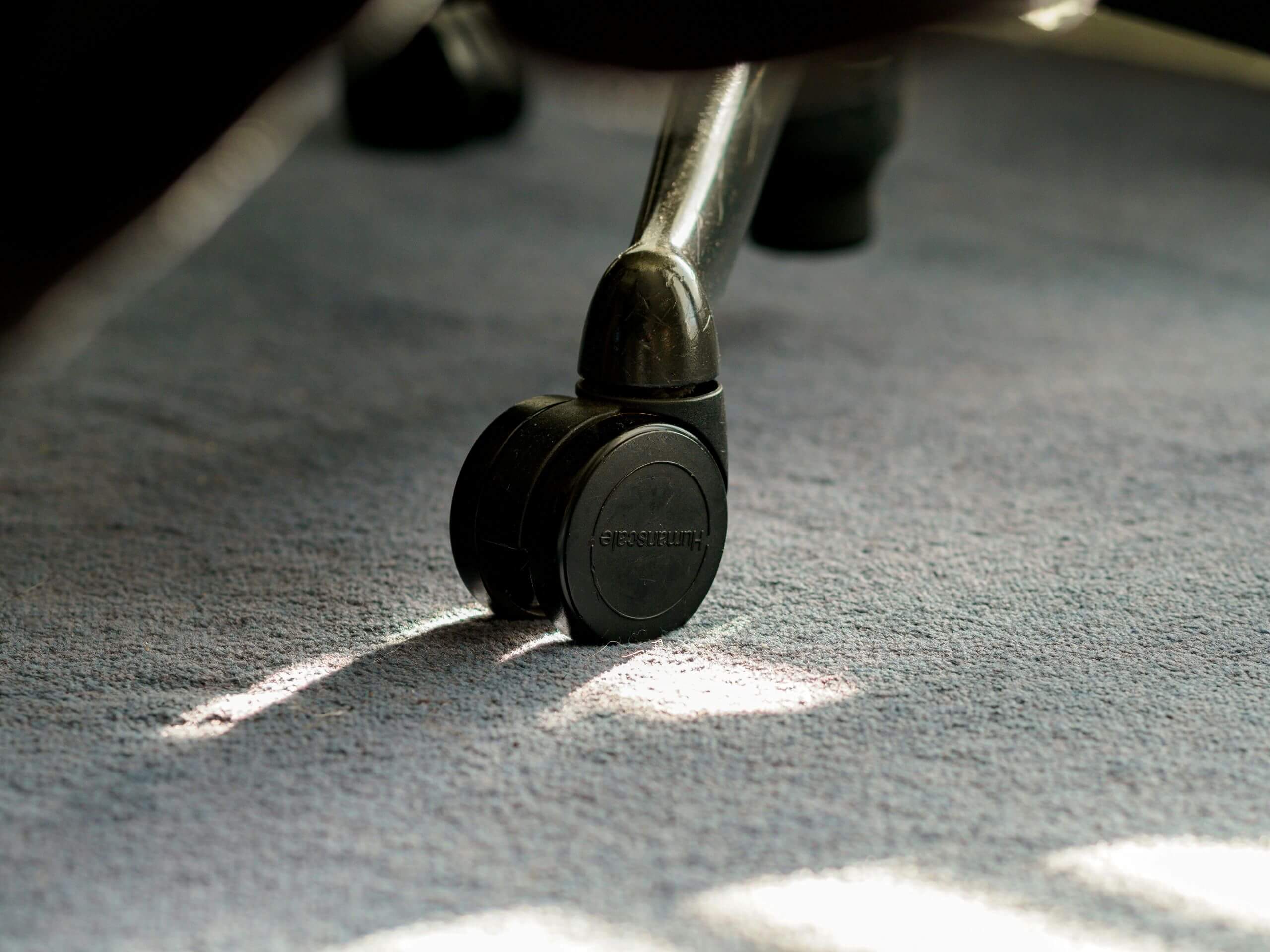 Close-up of a black plastic office chair wheel resting on a blue carpet, sunlight casting shadows—ideal for illustrating diy carpet care and smart flooring maintenance tips.