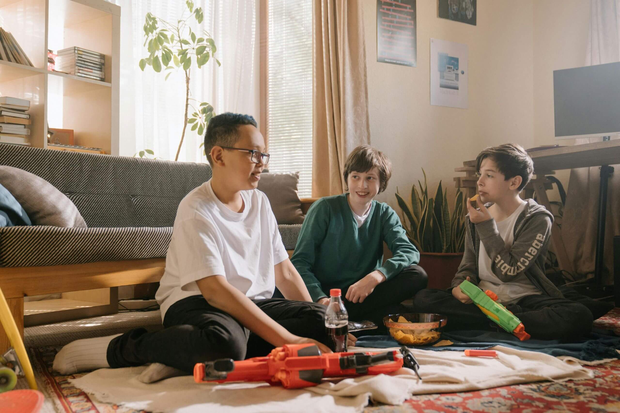 Three boys sit on the floor indoors, talking and smiling with toy blasters, snacks, and soda nearby. As sunlight fills the room with plants and a couch, they seem unaware of 5 carpet stains you should never try to remove on your own.