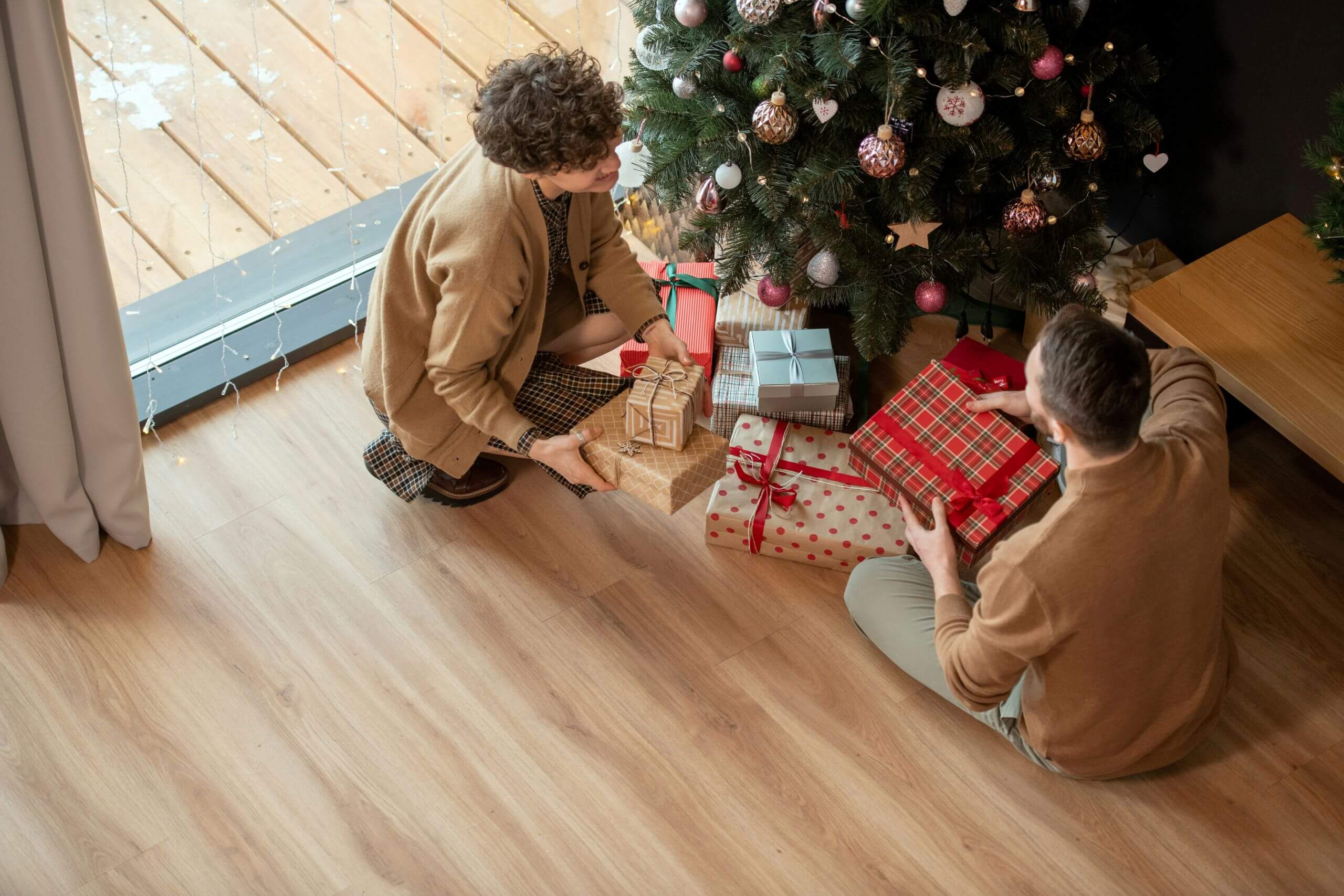 Two people sit on the floor near a decorated Christmas tree, arranging wrapped presents beneath it. The festive scene, bathed in natural light, hints at how cold weather affects hardwood floors during the cozy holiday season.