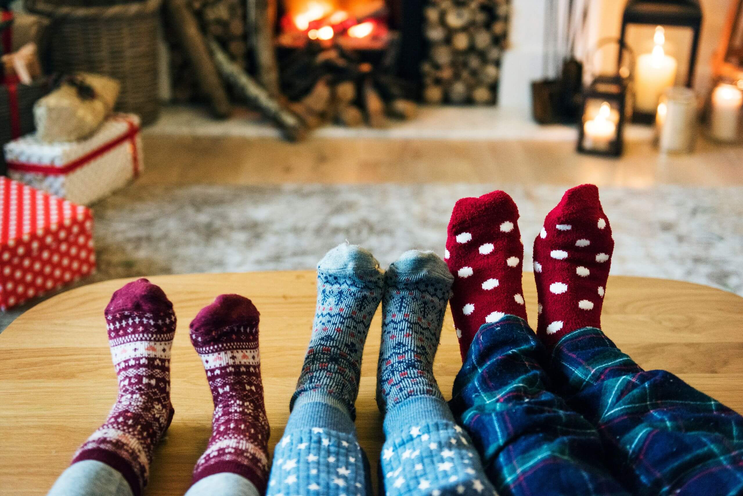 Four people wearing colorful patterned socks rest their feet on a wooden table in front of a cozy fireplace. Wrapped gifts and candles set the mood, making it the perfect scene to inspire your winter floor care routine.