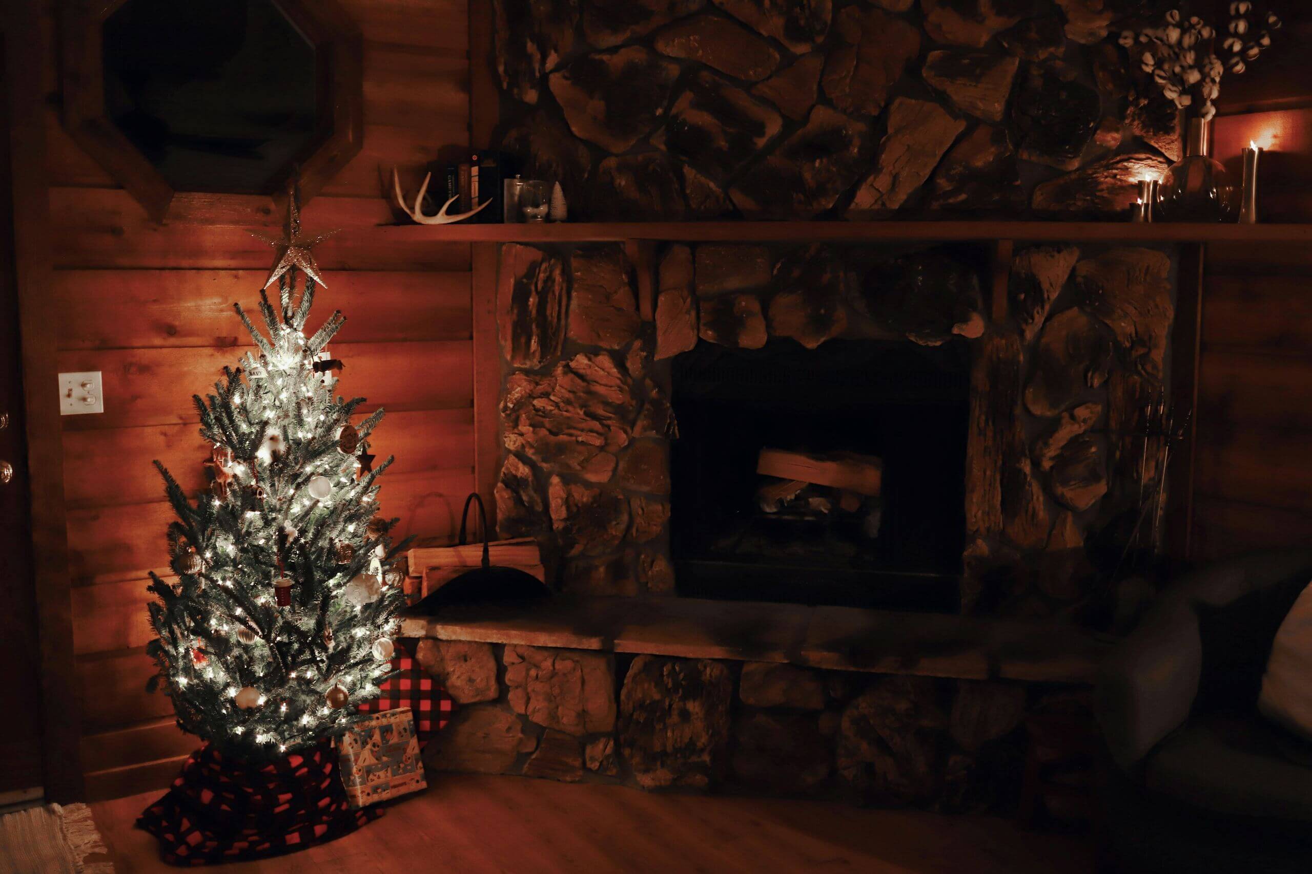 A small, lit Christmas tree stands next to a stone fireplace in a cozy, dimly lit wooden room, decorated with candles and rustic decor. Hardwood floor protection ensures the scene stays warm, festive, and worry-free.