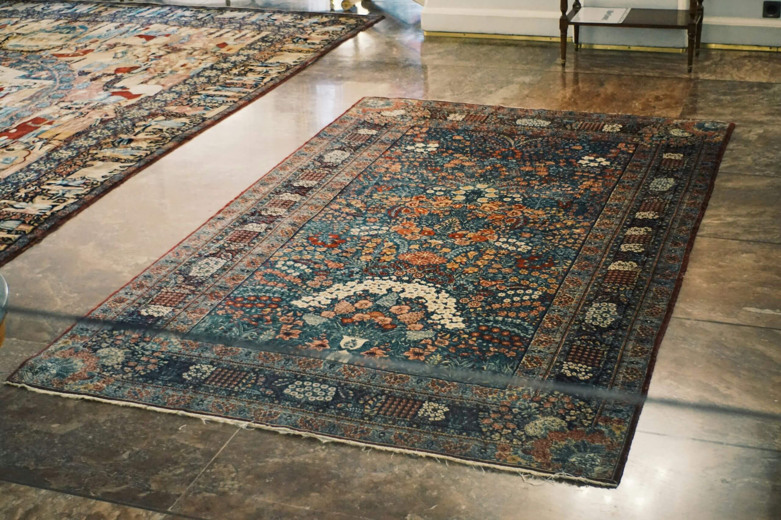 A rectangular, ornately patterned oriental rug with floral and geometric designs in blue, red, and cream colors lies on a polished marble floor in a well-lit room, looking fresh after post-holiday carpet cleaning.