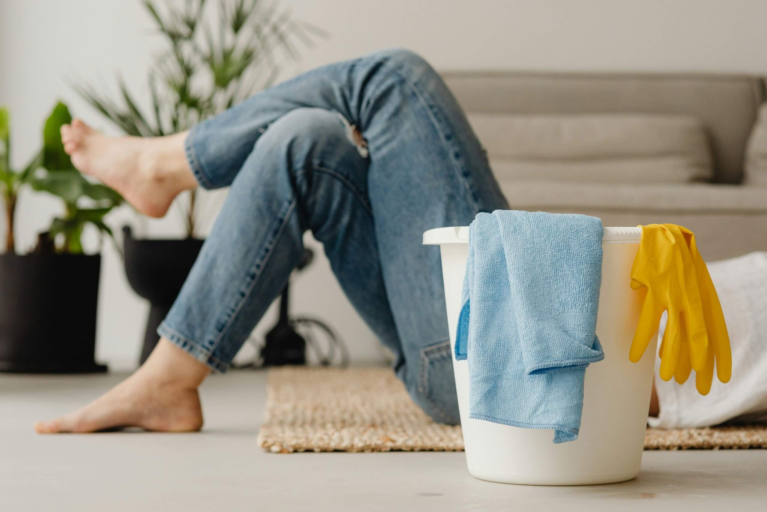A person in jeans rests on the floor near a sofa, legs stretched out after spring cleaning. In the foreground, a white bucket holds a blue cloth and yellow cleaning gloves. Houseplants are visible in the background.