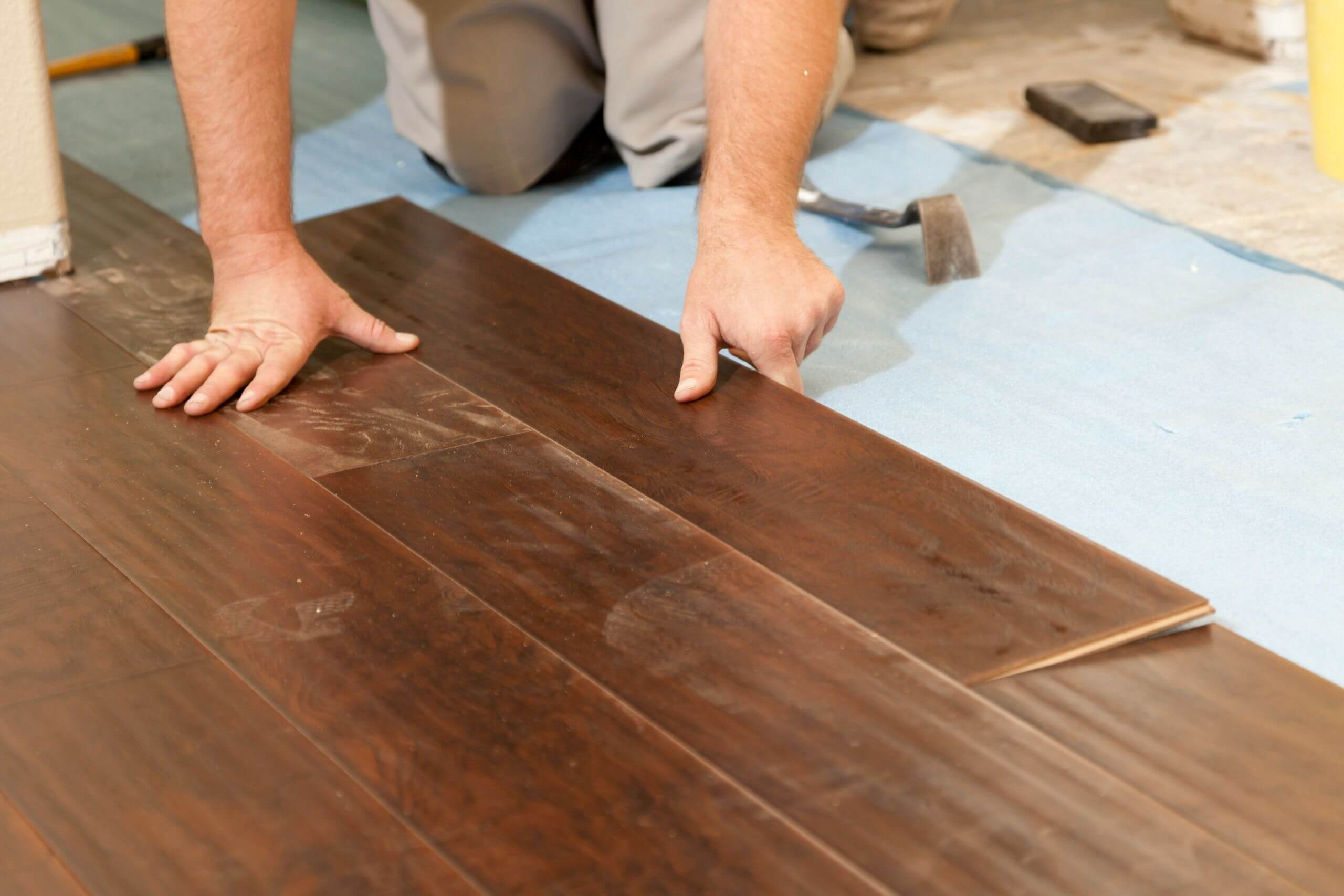 A person is installing dark wood flooring to replace your floors, fitting a plank into place on a blue underlayment. Tools and flooring materials are visible nearby.