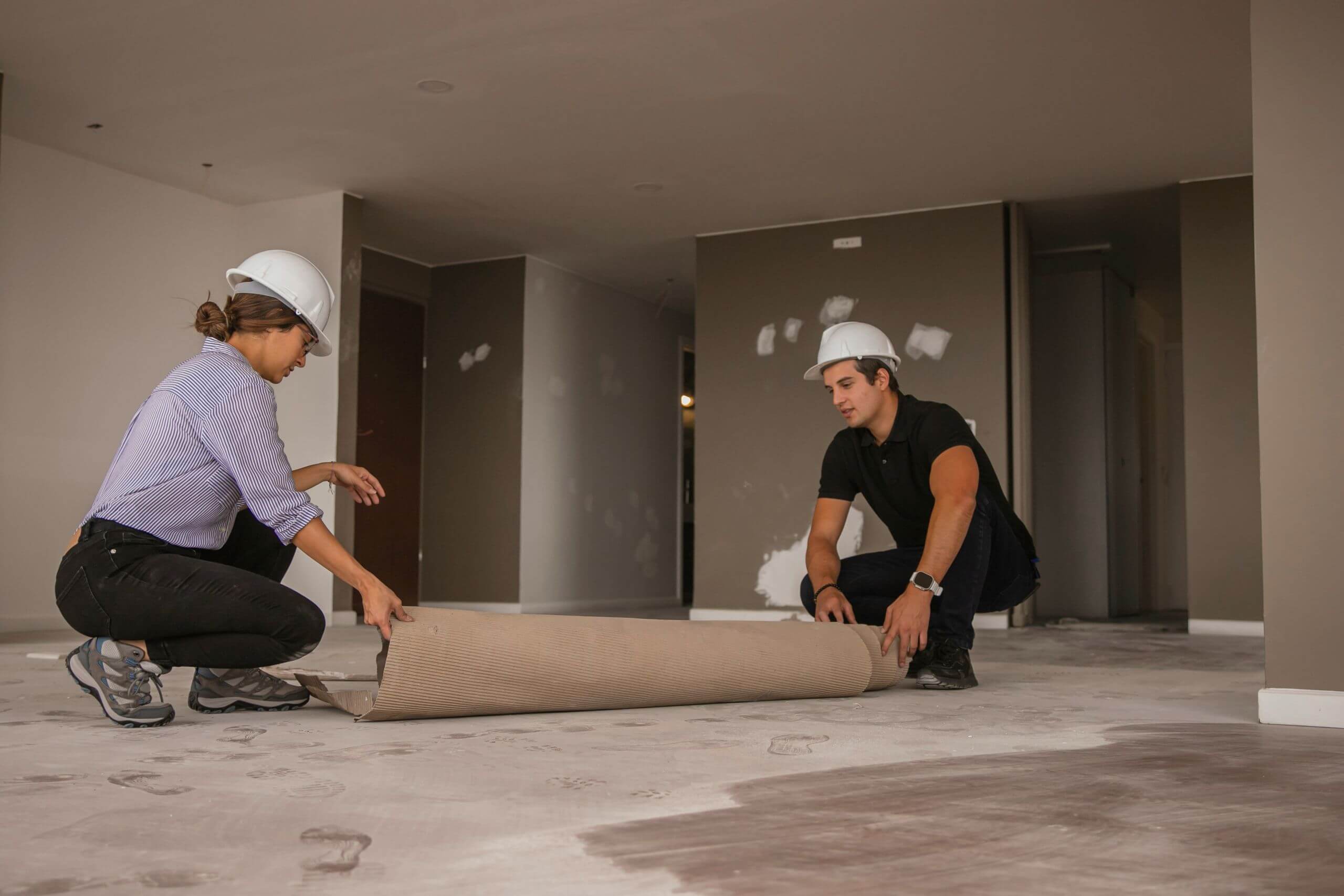 Two construction workers in hard hats unroll a carpet in an unfinished room, showing a flooring maintenance.