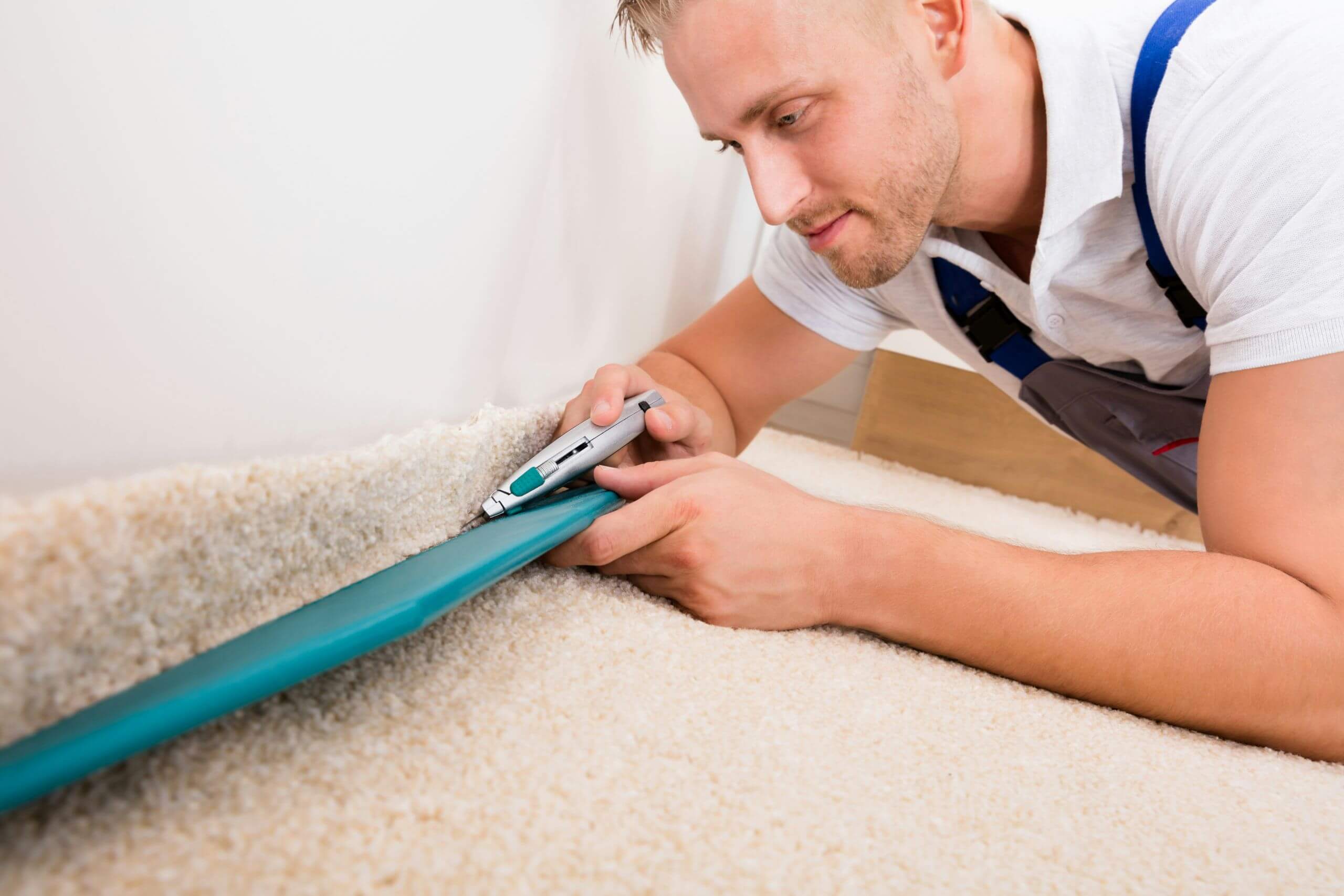 A carpet installer doing Carpet Removal and Floor Installation. He cuts a piece of carpet with a utility knife, using a blue guide to ensure a straight edge.