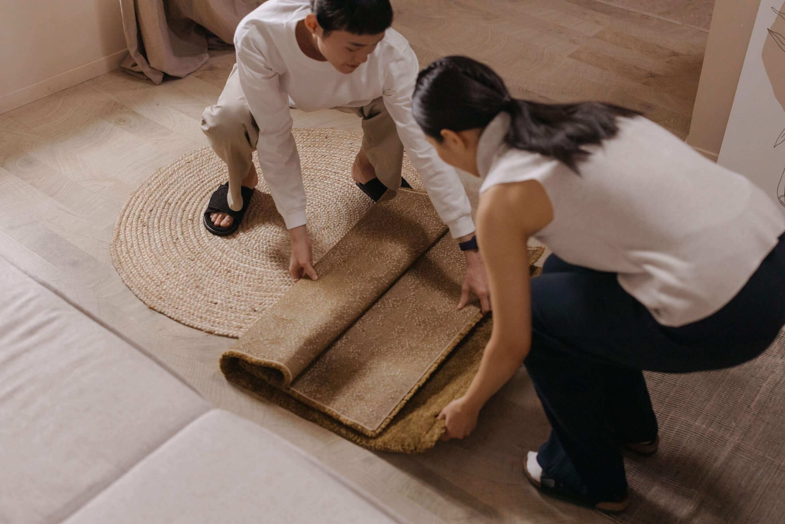 Two people are kneeling on the floor, working together to roll up a beige carpet on top of a round woven mat in a cozy indoor setting, showing one of the carpet maintenance tips