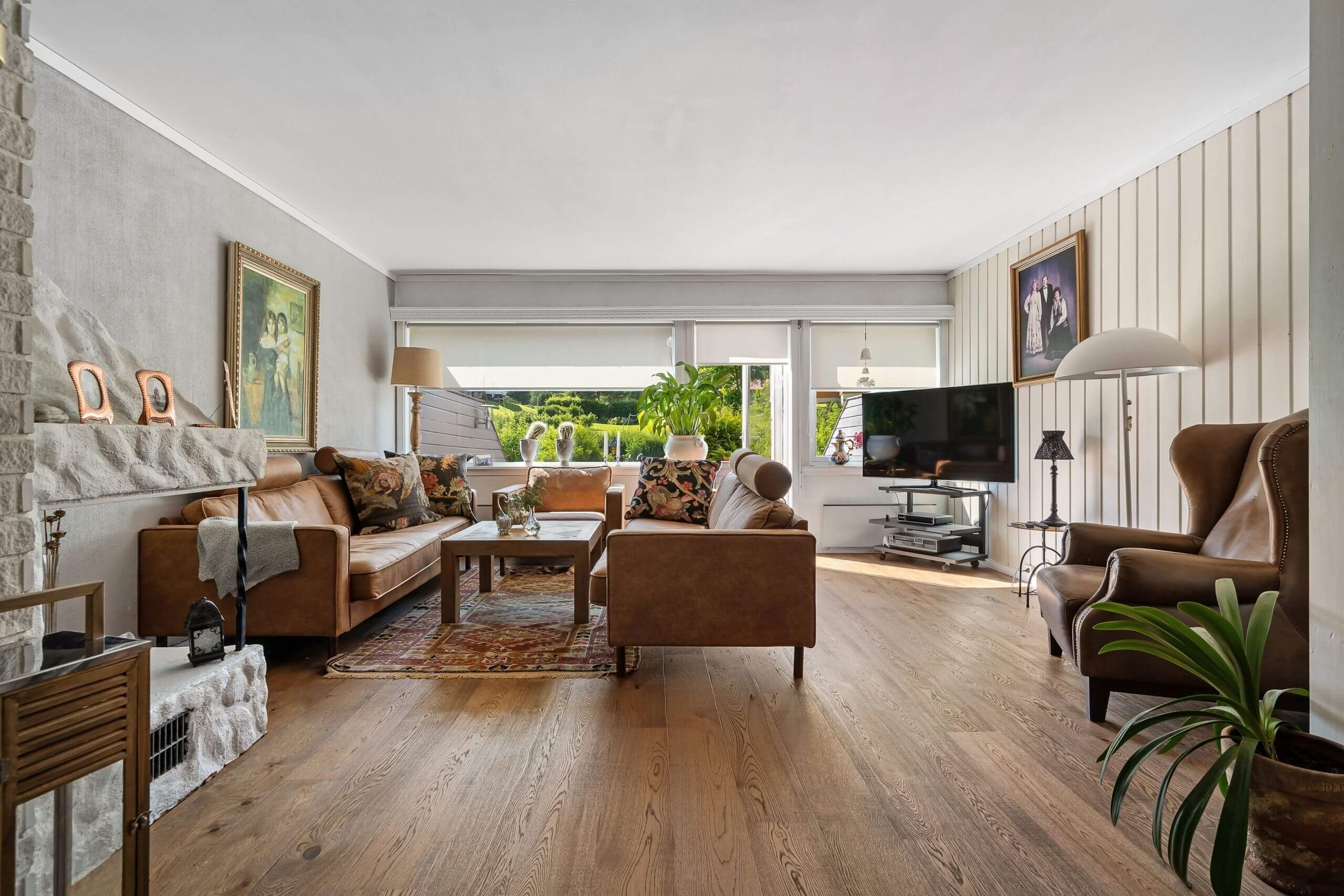 A spacious living room with flawless wooden floors—no flooring failure here—brown leather sofas, armchairs, a white stone fireplace, large windows framing lush outdoor views, paintings on the walls, and a TV on a stand in the corner.
