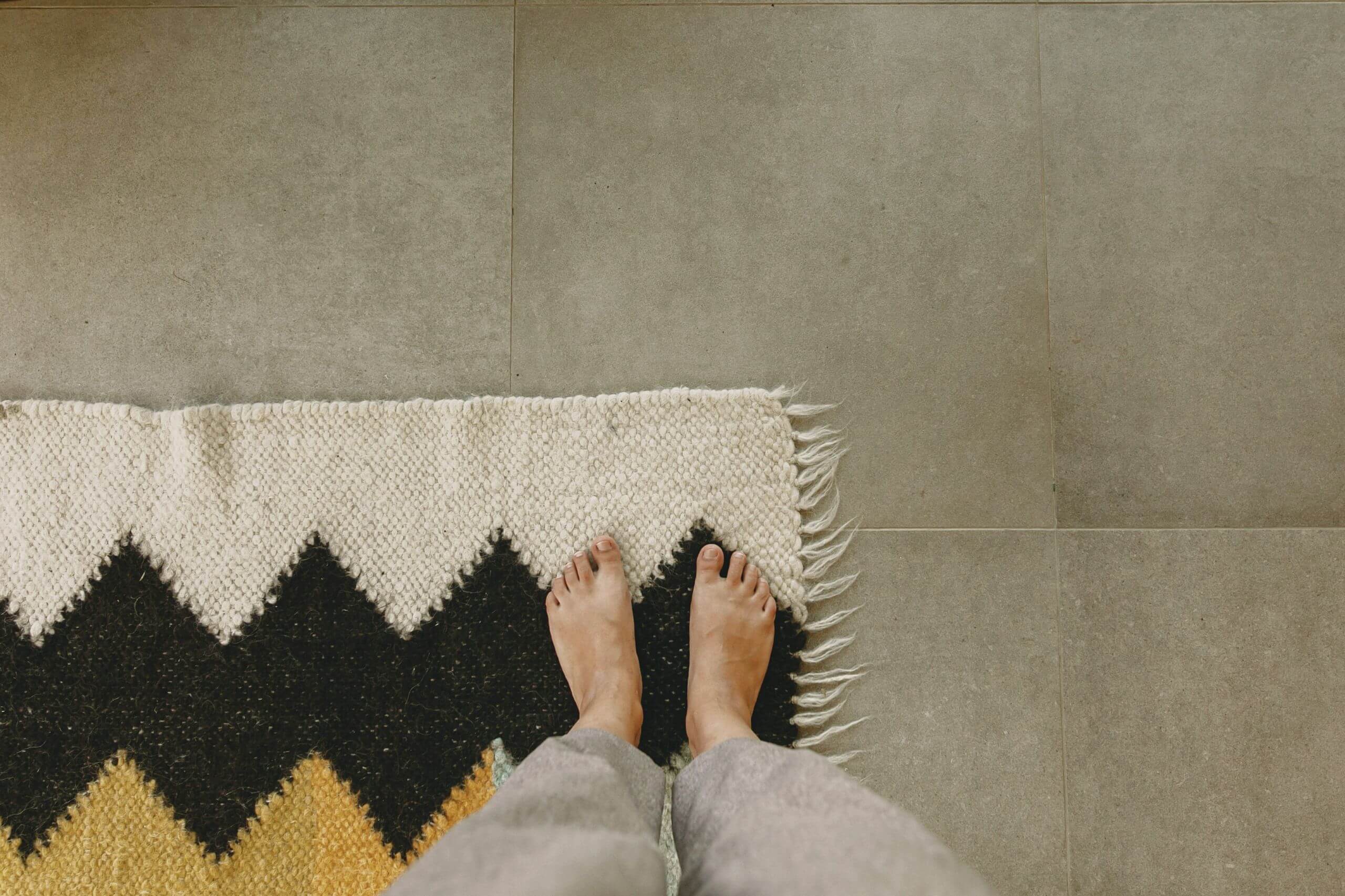 Person standing barefoot on a textured rug with black and yellow zigzag patterns, next to gray floor tiles, showing ideal area rugs for hardwood floors