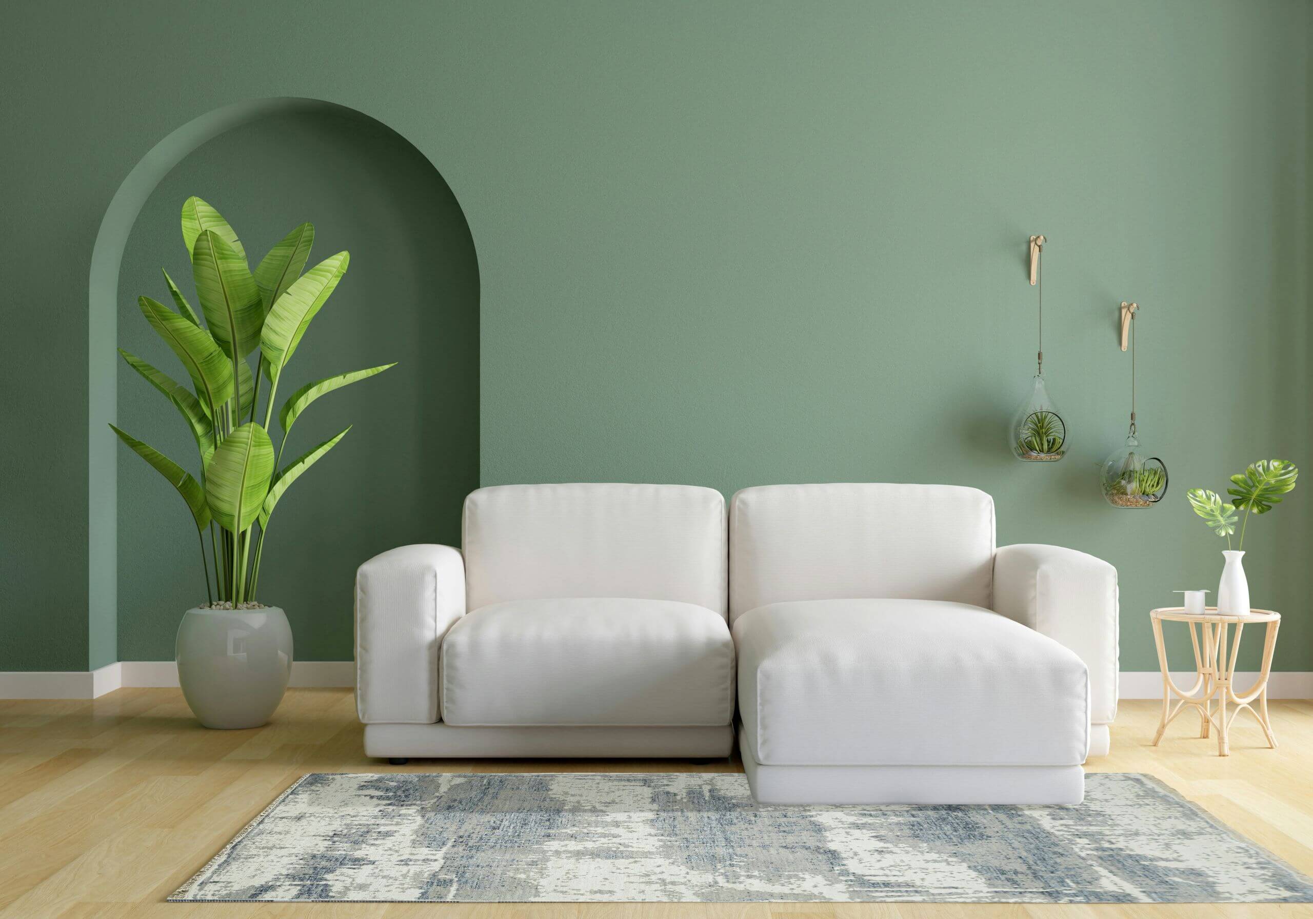 A modern living room with a white sofa, green wall, potted plant, two hanging glass planters, a small side table with a plant, and a blue patterned rug on a light wood floor.