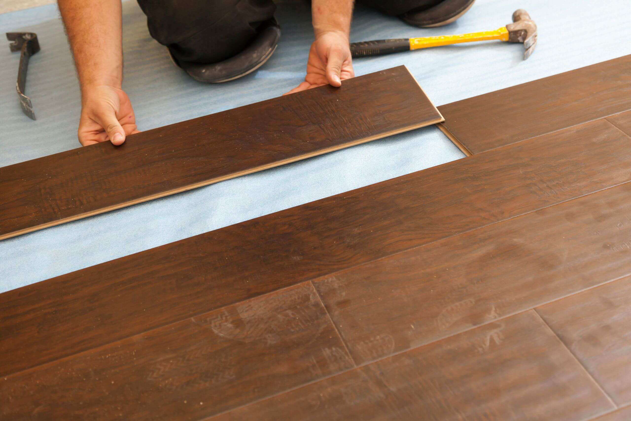 A person installs dark wood laminate flooring on a blue underlayment, aligning a plank with the others, showing how he knows how to choose the best flooring for home. A hammer and pry bar are nearby.