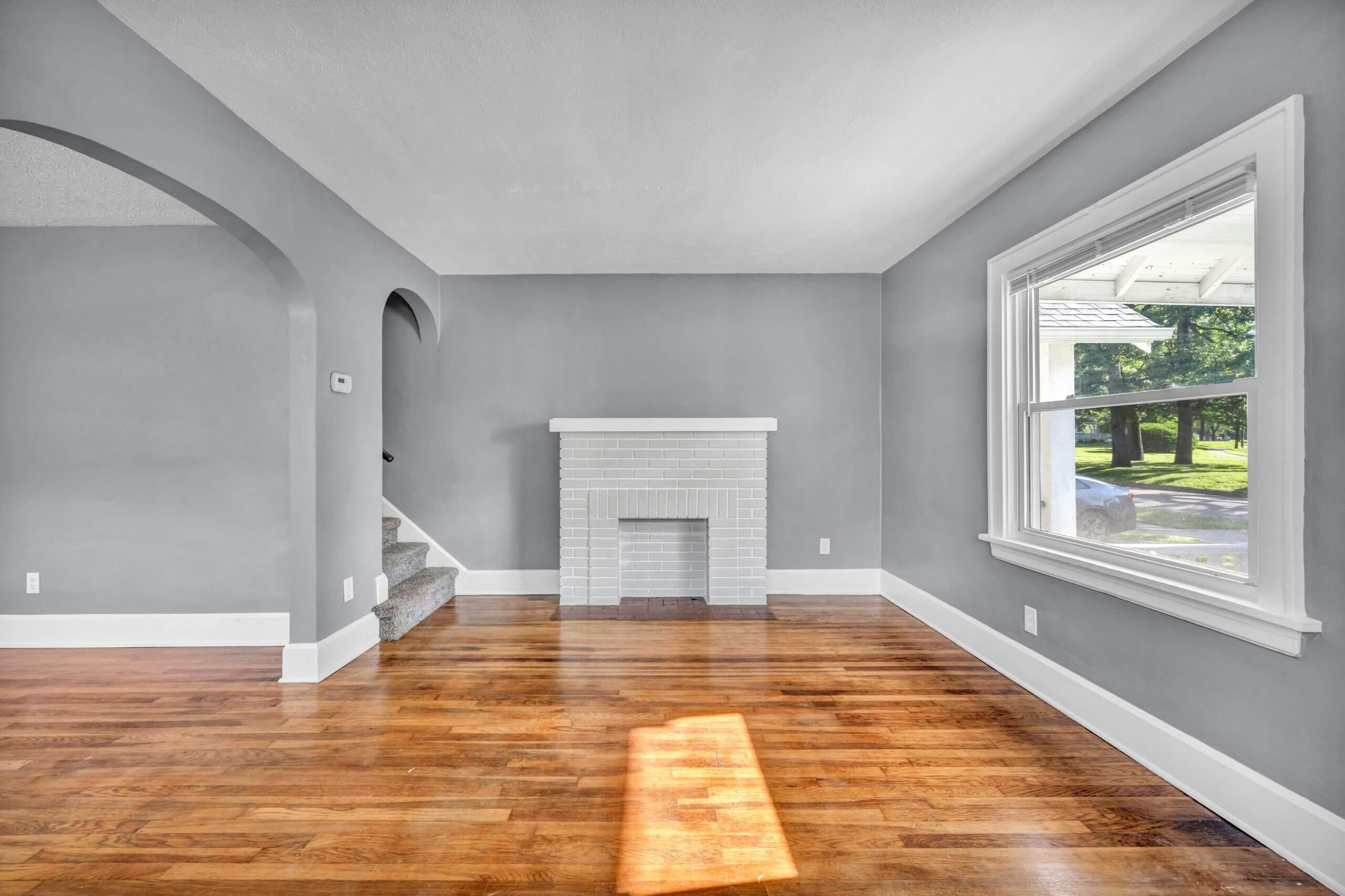 Bright living room with gray walls, white trim, hardwood floors, a white brick fireplace, carpeted stairs, an arched doorway, and a large window letting in natural light.