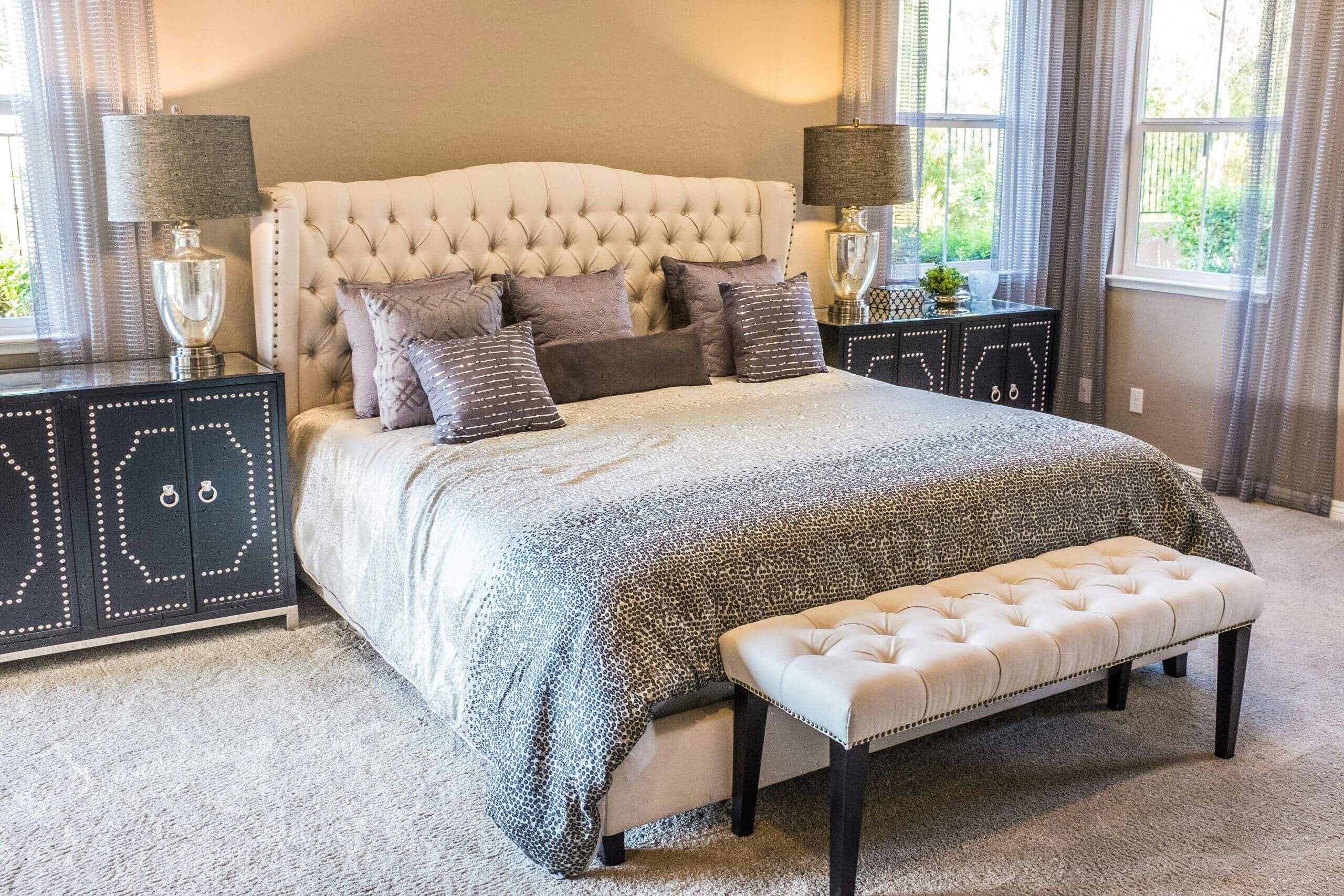 A bedroom with a tufted cream headboard, a gray patterned duvet, two dark bedside cabinets with silver trim, and a tufted cream bench at the foot of the bed.